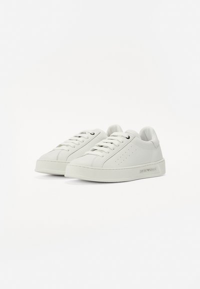 Hvite lær sneakers med rund tå, flate snører, perforeringer på siden, og en teksturert såle. Har merking på utsiden.