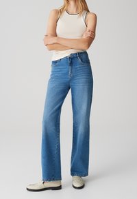 Camiseta sin mangas beige claro con ribete negro, combinada con vaqueros de pierna ancha azules y mocasines blancos. Tejido suave, corte relajado y textura clásica de denim.