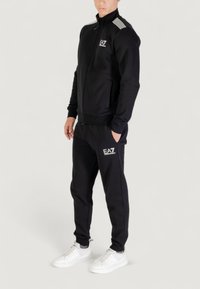 Chaqueta negra con cremallera y logo blanco en el pecho, acentos a rayas en los hombros, combinada con pantalones a juego y zapatillas blancas.