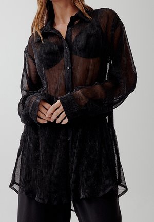 Kobieta nosząca prześwitującą czarną koszulę z długim rękawem i guzikami na czarnym biustonoszu, z rękoma luźno złożonymi na talii.