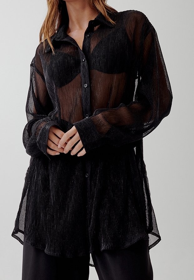 Femme portant une chemise noire transparente à manches longues boutonnée sur un soutien-gorge noir, les mains légèrement jointes à la taille.