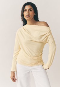 Haut jaune à manches longues avec devant drapé et design épaules dénudées, fabriqué en tissu doux. Associé à un pantalon blanc, avec des poignets ajustés.