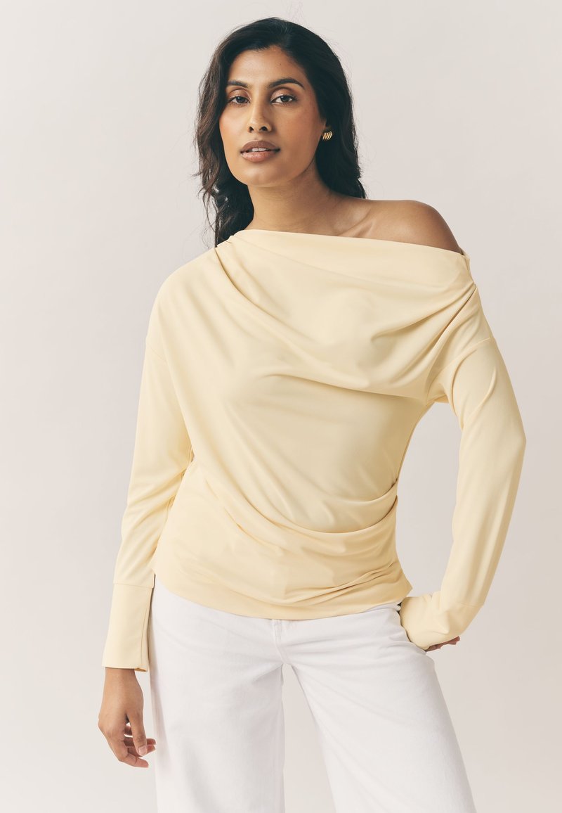 Haut jaune à manches longues avec devant drapé et design épaules dénudées, fabriqué en tissu doux. Associé à un pantalon blanc, avec des poignets ajustés.