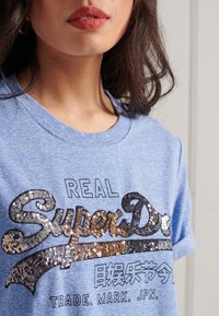Camiseta de algodón azul con cuello redondo y mangas cortas, adornada con un logo de "SuperDry" con lentejuelas.