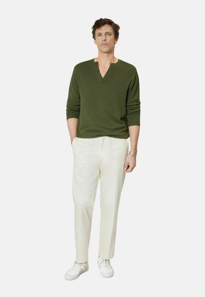 Hombre con suéter de cuello en V color verde oliva, pantalones blancos, zapatillas blancas, de pie con una mano en el bolsillo contra un fondo liso.