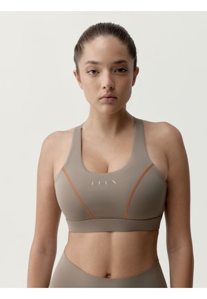 Femme aux cheveux attachés portant un brassard de sport taupe et un legging assorti, debout sur un fond clair uni.