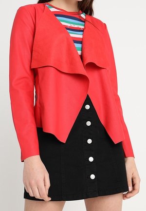 Vrouw die een felrode jas met open voorkant draagt over een gestreept shirt, gecombineerd met een zwarte rok met knopen.