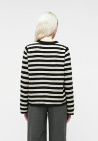 Osoba z długimi jasnymi włosami nosi czarno-biały sweter w poziome paski i ciemnoszare spodnie, stojąc na tle jednolitego jasnego tła.