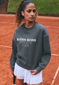 Sudadera gris con "BJÖRN BORG ATHLETIC CLUB" en texto blanco, combinada con una falda blanca con pliegues. Textura suave, mangas largas y corte relajado.