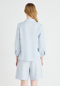 Top in lino blu chiaro con maniche lunghe e arricciate, abbinato a pantaloni corti coordinati, che mettono in mostra un fit rilassato e un design minimalista.