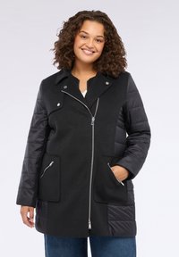 Abrigo negro con sección superior texturizada y mangas acolchadas brillantes. Incluye un cierre de cremallera, bolsillos y un diseño de cuello con muesca.