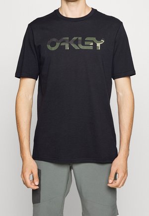 Mann trägt schwarzes Oakley-T-Shirt mit grünem Camouflagelogo und graue Hose mit schwarzem Patch am Oberschenkel, steht vor einfarbigem Hintergrund.