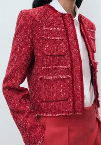 Roter Tweed-Jacke mit fransigen Kanten, die über vier Taschen und ein kurzes Design verfügt. Kombiniert mit einem weißen Hemd mit Knöpfen und roten Hosen.