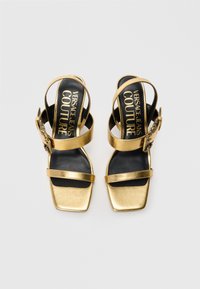 Gold high-heeled sandals with a square toe, featuring two wide straps and an ankle strap with a decorative buckle, black interior lining.