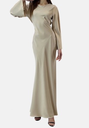 Femme portant une longue robe en satin beige avec de longues manches larges et des sandales noires à talons hauts ouvertes à l'avant, posant avec une main près du visage.