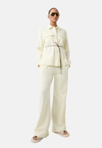 Chemise crème à boutons avec deux poches, associée à un pantalon large. Fini avec une cravate marron à la taille et des sandales beiges volumineuses.