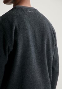 Persona che indossa un maglione grigio scuro con texture, con un piccolo marchio ricamato vicino al colletto sul retro, su uno sfondo chiaro.