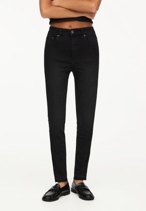Mujer con vaqueros pitillo negros de talle alto combinados con mocasines negros, de pie con los brazos cruzados sobre un fondo blanco.