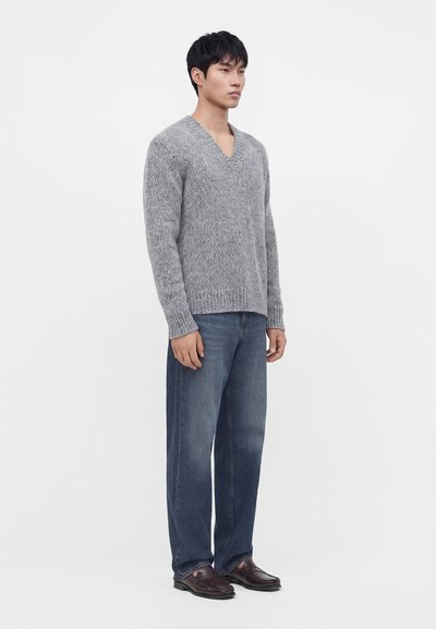 Jeune homme debout portant un pull gris à col en V, un jean bleu ample et des chaussures en cuir brun foncé, sur un fond clair uni.