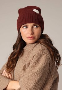 Femme aux longs cheveux bruns portant un bonnet tricoté bordeaux avec un cœur blanc et une veste beige texturée, regardant sur le côté les bras croisés.