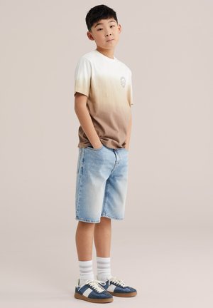 Berniukas stovintis su rankomis kišenėse, dėvintis smėlio spalvos ombre marškinėlį, šviesiai mėlynas džinsines šortus, baltas kojines ir mėlynas sportbačius.