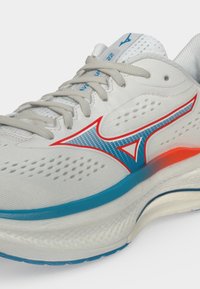 Chaussure de course blanche avec lacets gris, logo Mizuno bleu et rouge, côté perforé, et détail de semelle bleu et orange.