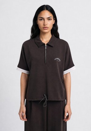 Woman with long dark hair wearing a dark brown short-sleeve top with a zipper and drawstring, standing against a plain light background.