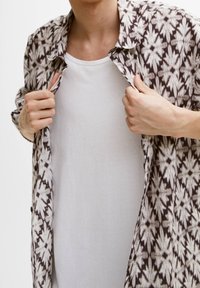 Camisa de patrón marrón y blanco con una tela ligera, abierta por delante, sobre una camiseta blanca básica, que muestra un ajuste relajado.