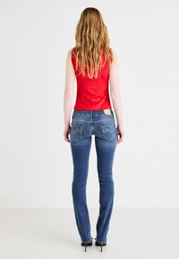 Modre jeans hlače prilegajoče kroja z petimi žepi in majhno usnjeno nalepko na zadnji strani. Narejene z rdečim brezrokavnikom.