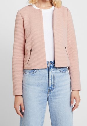 Frau trägt eine hellrosa strukturierte Jacke über einem weißen Hemd und hellblaue Jeans mit hoher Taille, steht mit entspannten Armen an den Seiten.