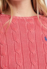 Pull en maille torsadée rose avec un col rond, orné d'un patch logo et de motifs texturés en relief sur tout le tissu.