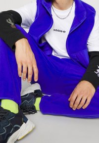 Lila fleeceväst och byxset, vit t-shirt med svart text, svart långärmad tröja med kaktustryck, svarta sneakers med gula sulor.