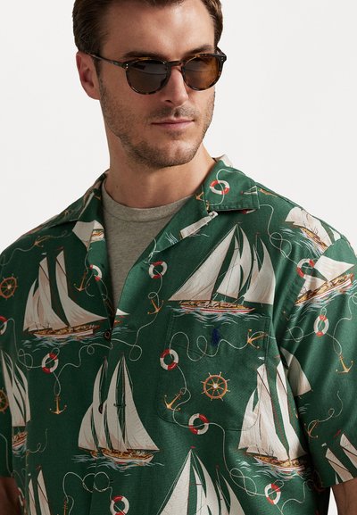 Camisa verde de manga curta com um padrão náutico de barcos à vela, âncoras e boias de salvamento. O tecido parece leve e suave.
