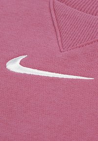 Roosa kootud fliis, V-kaelusega ja soonikkoelise kraega. Esiküljel valge Tikitud Nike Swoosh logo. Pehme tekstuur.