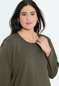 Chemise à manches longues vert olive avec un col rond et une coupe décontractée, fabriquée en tissu doux. Présente des détails cousus le long de l'ourlet.