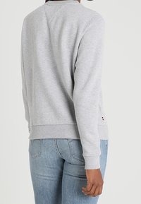 Persona che indossa una felpa a maniche lunghe grigio chiaro e jeans azzurri, mostrata di spalle su uno sfondo semplice.