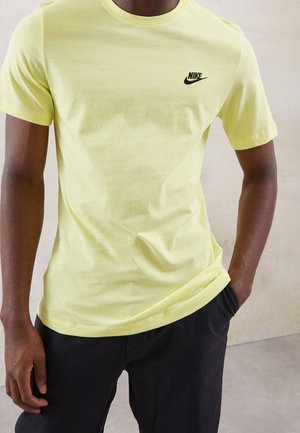 Mann trägt ein hellgelbes Nike-T-Shirt mit schwarzem Logo auf der linken Brust, schwarze Hose, steht mit einer Hand in der Tasche.