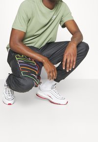 Man som hukar i en ljusgrön Nike-t-shirt, svarta byxor med färgglatt Nike-logotyp, vita strumpor och vita Nike-sneakers med röda detaljer.