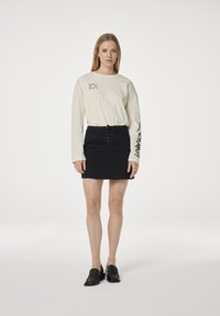 Jonge vrouw staat in een crèmekleurig shirt met lange mouwen en groene tekst, een zwarte minirok met veters en zwarte instapschoenen op een witte achtergrond.