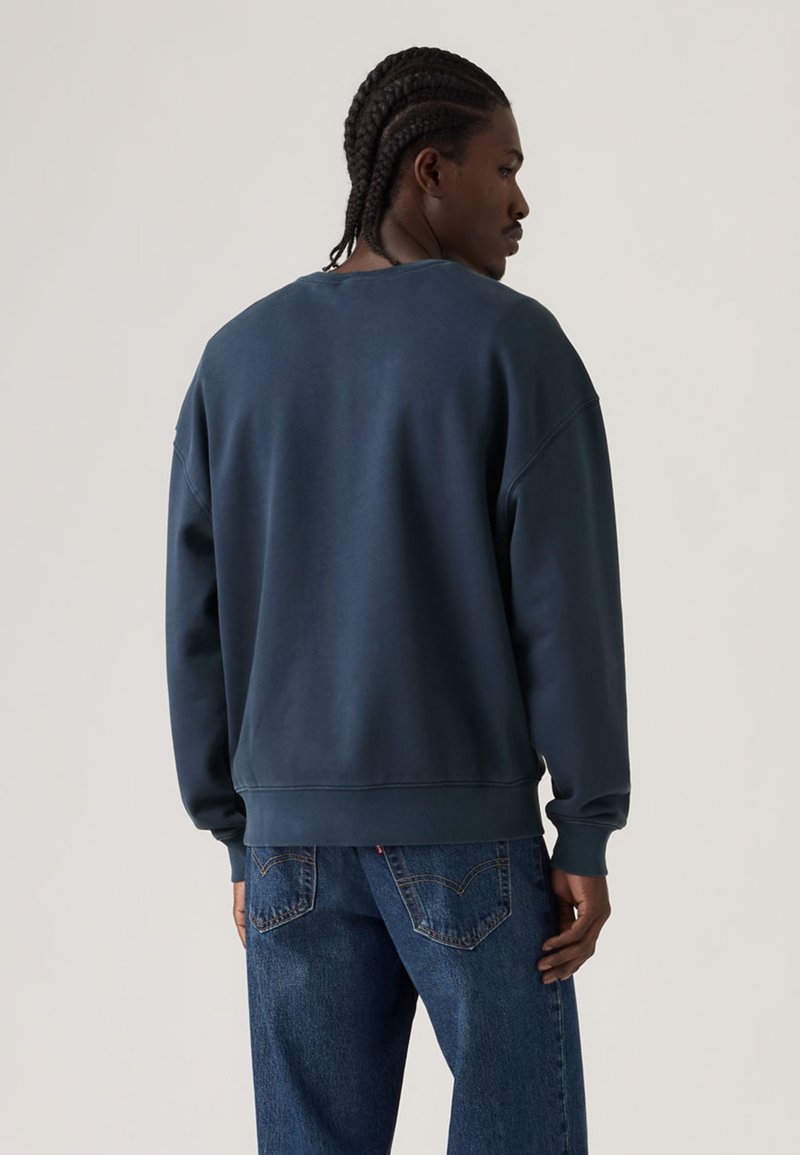 Felpa blu scuro con scollo rotondo e spalle scese, realizzata in tessuto morbido. Presenta polsini e orlo a coste. Indossata con jeans blu.