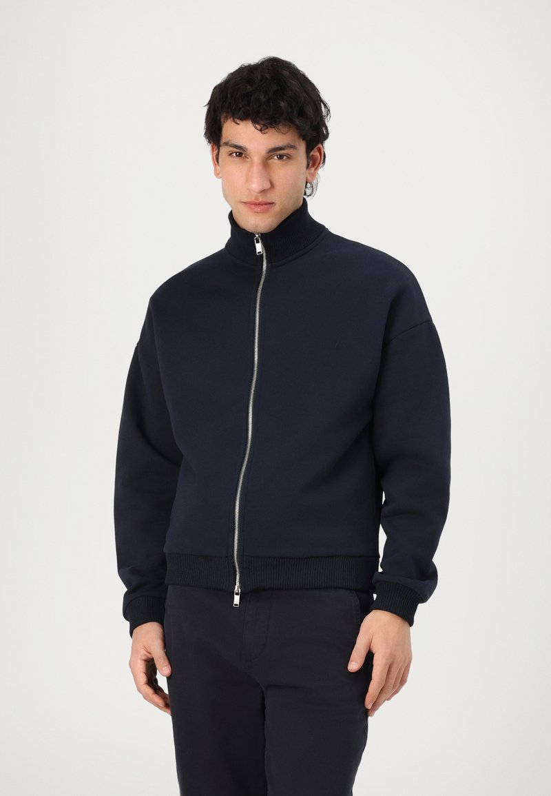 Marineblå zip-up hettegenser med høy krage, ribbestrikkede mansjetter og liv; laget av mykt stoff; med sølvglidelås og minimalistisk design.