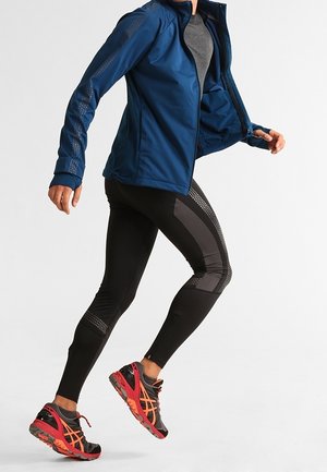 Persona vestida con chaqueta azul, camiseta gris, mallas deportivas negras y zapatillas de correr rojas en medio de un zancada frente a un fondo liso.