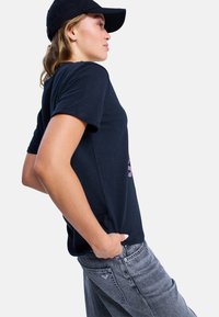 Young woman in black cap, navy blue t-shirt, and gray jeans posing sideways with hands in pockets against white background.