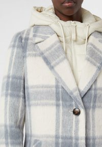 Cappotto in misto lana a quadri con motivo grigio chiaro e crema, caratterizzato da un colletto a V e una chiusura a singolo bottone. Fodera con uno strato interno trapuntato beige.