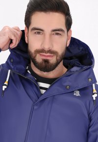 Granatowa wodoodporna kurtka z zamkiem błyskawicznym, kapturem ze sznurkiem i logo. Nosi ją osoba założona na czarno-biało paskowaną koszulę.