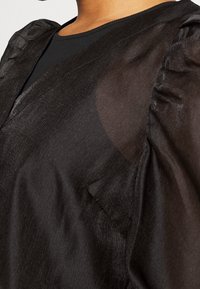 Top nero con tessuto trasparente e testurizzato, con maniche arricciate e scollatura a V. Il materiale è leggero e leggermente lucido.