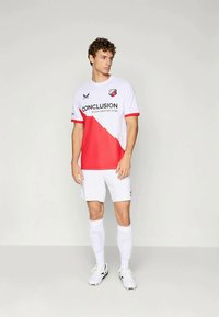 Maglia da calcio bianca con dettagli rossi, caratterizzata da un design diagonale e testo stampato. Abbinata a pantaloncini e calzettoni bianchi; calzature atletiche.