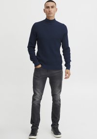 Mornarsko pleteno pulover z visoko ovratnico in dolgimi rokavi, z rebrastimi manšetami in robom, v kombinaciji s sivo usklajenimi jeansi in črnimi supergami.