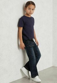 Jūras zilas kokvilnas T-kreklas kombinēts ar tumšiem džinsiem ar taisnu piegriezumu, ar šuvju akcentiem un baltām sneaker kurpēm. Bērns balstās pret sienu.