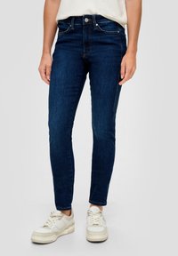 Jeans skinny azul oscuro de denim, con un corte de talle alto, bolsillos frontales y detalles de costura sutiles. Combinados con zapatillas blancas.